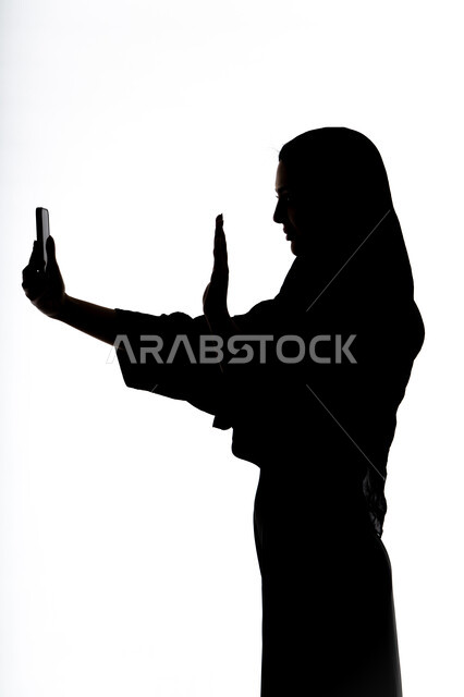 Using a mobile phone, communicating with family and friends, taking souvenir selfies, using modern technology devices, conducting a live broadcast, making a phone call, silhouette of an Arab Gulf Emirati woman, social media, white background