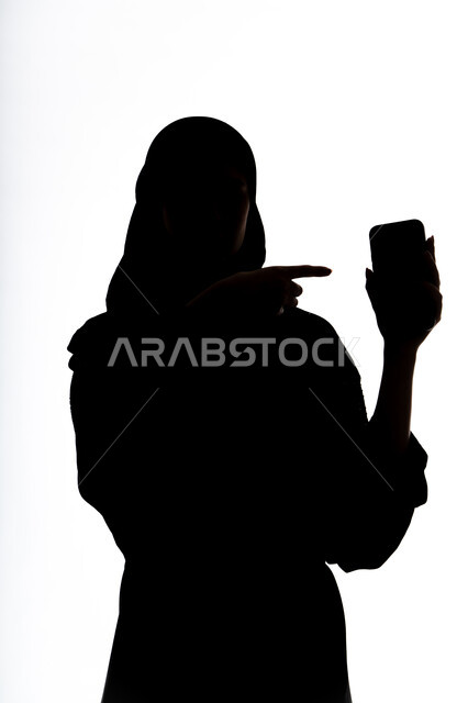الاشاره الى شاشه الجوال، التواصل مع الاهل والاصدقاء، التقاط صور سيلفي تذكاريه ،إستخدام اجهزة تقنية حديثة ، اجراء بث مباشر ،  اجراء مكالمه هاتفيه ،صورة ظلية لمرأة عربية خليجية اماراتيه ،وسائل التواصل الإجتماعي ، خلفية بيضاء