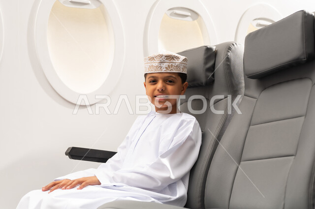 Tourist trips in the Sultanate of Oman, a Gulf Arab Omani boy wearing a dishdasha and a kumma sits in the airplane cabin by the window looking at the camera with gestures of joy and happiness, the concept of tourism and travel, Oman Air.