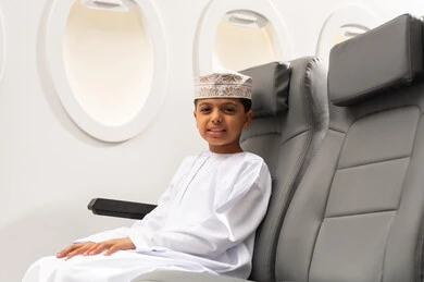 الرحلات السياحية في سلطنه عمان, ولد عربي خليجي عماني يرتدي الدشداشة و الكمة يجلس في مقصورة الطائرة بجانب النافذة ينظر الى الكاميرا بإيماءات الفرح والسرور, مفهوم السياحة والسفر، خطوط الطيران الجوية العمانية