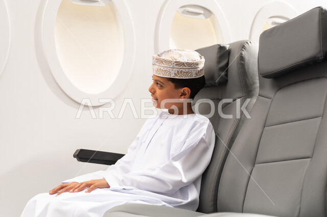 Getting ready for an enjoyable summer vacation, a tourist trip with Oman Air, looking out the airplane window, an Arab Gulf Omani child wearing a dishdasha and a kumma sitting in the airplane seat.