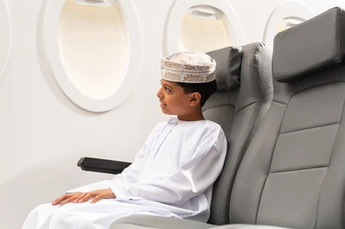 Getting ready for an enjoyable summer vacation, a tourist trip with Oman Air, looking out the airplane window, an Arab Gulf Omani child wearing a dishdasha and a kumma sitting in the airplane seat.