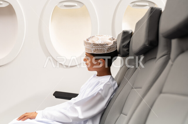رحلة سياحية من خلال خطوط الطيران العماني, النظر من نافذة الطيارة، طفل مسافر عربي خليجي عماني يرتدي الدشداشة و الكمه يجلس في مقعد الطائرة، الاستعداد لقضاء عطلة صيفية ممتعة
