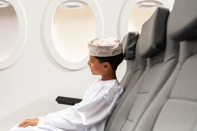 A tourist trip through Oman Air, looking out the airplane window, an Arab Gulf Omani child wearing a dishdasha and kumma sitting in the airplane seat, getting ready to enjoy a fun summer vacation.