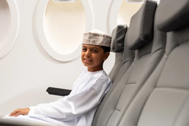 ولد عربي خليجي عماني يرتدي الدشداشة و الكمة يجلس في مقصورة الطائرة بجانب النافذة ينظر الى الكاميرا بإيماءات الفرح والسرور, مفهوم السياحة والسفر، خطوط الطيران الجوية العمانية، الرحلات السياحية في سلطنه عمان