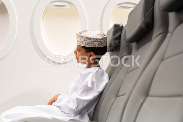 النظر من نافذة الطيارة، طفل مسافر عربي خليجي عماني يرتدي الدشداشة و الكمه يجلس في مقعد الطائرة، الاستعداد لقضاء عطلة صيفية ممتعة، رحلة سياحية من خلال خطوط الطيران العماني