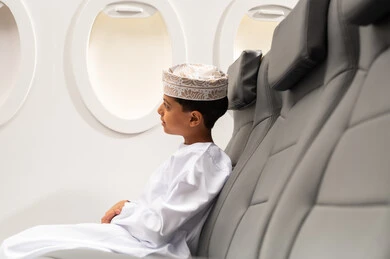 النظر من نافذة الطيارة، طفل مسافر عربي خليجي عماني يرتدي الدشداشة و الكمه يجلس في مقعد الطائرة، الاستعداد لقضاء عطلة صيفية ممتعة، رحلة سياحية من خلال خطوط الطيران العماني