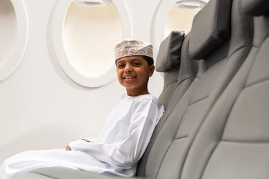 مفهوم السياحة والسفر، خطوط الطيران الجوية العمانية,ولد عربي خليجي عماني يرتدي الدشداشة و الكمة يجلس في مقصورة الطائرة بجانب النافذة ينظر الى الكاميرا بإيماءات الفرح والسرور، الرحلات السياحية في سلطنه عمان