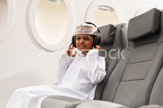 مفهوم الإصغاء والإستماع , الاستعداد لقضاء عطله صيفية ممتعه ، ولد عربي خليجي عماني يرتدي الدشداشة و الكمه يجلس داخل الطائرة يقوم بارتداء الهيدفون، الاستماع الى الموسيقى عبر سماعات الاذن