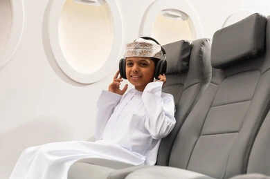 The concept of listening and hearing, preparing to spend an enjoyable summer vacation, an Arab Gulf Omani boy wearing a dishdasha and a kumma sitting inside the airplane putting on headphones, listening to music through earphones.