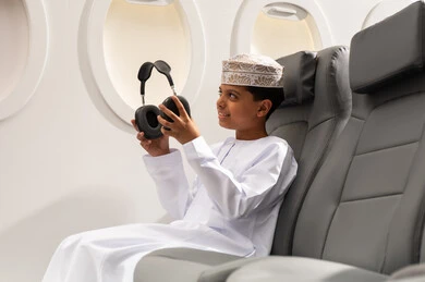 Integrating technology and modern techniques into daily life, the concept of tourism and travel, preparing for an enjoyable summer vacation, an Arab Gulf Omani boy wearing a dishdasha and kumma sitting inside the airplane putting on headphones, listening to music through earphones.