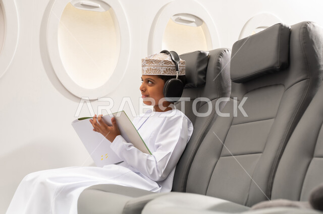 لعبة تلوين الرسومات, انشطة ترفيهية فنية ابداعية, ولد عربي خليجي عماني يرتدي الدشداشة و الكمة و السماعات يجلس على الكرسي داخل الطائرة يمسك القلم و الكراسة , خطوط الطيران العماني , مفهوم السياحة و السفر