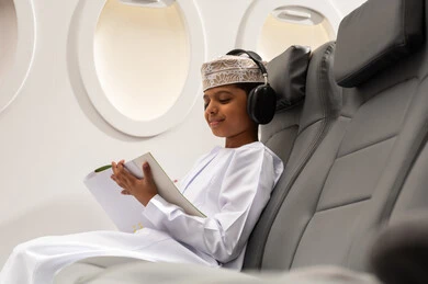 Creative artistic recreational activities, coloring game, an Arab Gulf Omani boy wearing a dishdasha and kumma and headphones sitting on a chair inside the airplane holding a pen and a drawing notebook, Oman Air, the concept of tourism and travel.