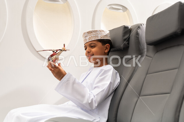 ايماءات وجه تدل على السعادة, قضاء وقت ممتع في الأجواء, رحلة سياحية من خلال خطوط الطيران العماني, صورة من الجانب لولد عربي خليجي عماني يرتدي الدشداشة و الكمه يجلس في مقعد الطائرة ويلعب بمجسم صغير لطائرة