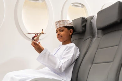 ايماءات وجه تدل على السعادة, قضاء وقت ممتع في الأجواء, رحلة سياحية من خلال خطوط الطيران العماني, صورة من الجانب لولد عربي خليجي عماني يرتدي الدشداشة و الكمه يجلس في مقعد الطائرة ويلعب بمجسم صغير لطائرة