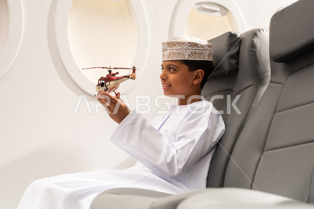 قضاء وقت ممتع في الأجواء, رحلة سياحية من خلال خطوط الطيران العماني, صورة من الجانب لولد عربي خليجي عماني يرتدي الدشداشة و الكمه يجلس في مقعد الطائرة ويلعب بمجسم صغير لطائرة، ايماءات وجه تدل على السعادة