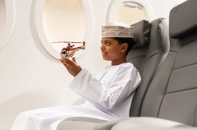 قضاء وقت ممتع في الأجواء, رحلة سياحية من خلال خطوط الطيران العماني, صورة من الجانب لولد عربي خليجي عماني يرتدي الدشداشة و الكمه يجلس في مقعد الطائرة ويلعب بمجسم صغير لطائرة، ايماءات وجه تدل على السعادة