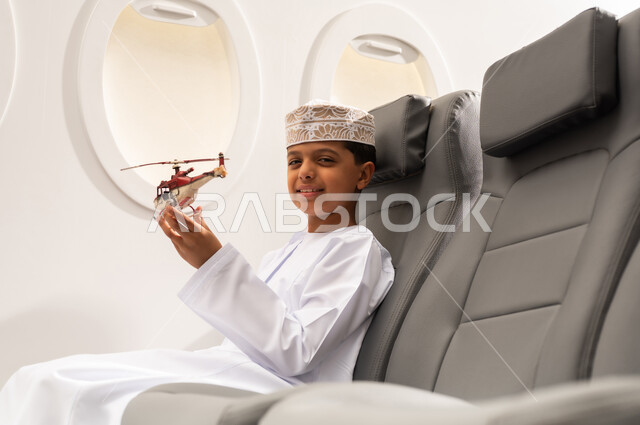 الاستعداد لقضاء عطلة صيفية ممتعة, رحلة سياحية من خلال خطوط الطيران العماني, صورة من الجانب لولد عربي خليجي عماني يرتدي الدشداشة و الكمه يجلس في مقعد الطائرة ويلعب بمجسم صغير لطائرة، ايماءات وجه تدل على السعادة