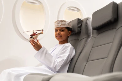 الاستعداد لقضاء عطلة صيفية ممتعة, رحلة سياحية من خلال خطوط الطيران العماني, صورة من الجانب لولد عربي خليجي عماني يرتدي الدشداشة و الكمه يجلس في مقعد الطائرة ويلعب بمجسم صغير لطائرة، ايماءات وجه تدل على السعادة