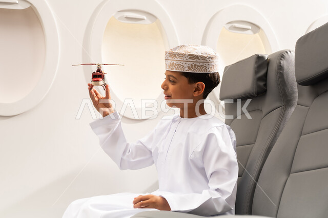 رحلة سياحية من خلال خطوط الطيران العماني, صورة من الجانب لولد عربي خليجي  عماني يرتدي الدشداشة و الكمه يجلس في مقعد الطائرة ويلعب بمجسم صغير لطائرة، ايماءات وجه تدل على السعادة، الاستعداد لقضاء عطلة صيفية ممتعة