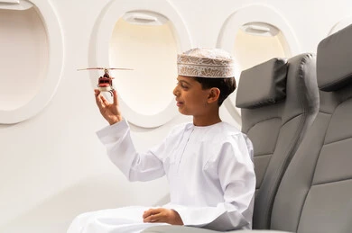 رحلة سياحية من خلال خطوط الطيران العماني, صورة من الجانب لولد عربي خليجي  عماني يرتدي الدشداشة و الكمه يجلس في مقعد الطائرة ويلعب بمجسم صغير لطائرة، ايماءات وجه تدل على السعادة، الاستعداد لقضاء عطلة صيفية ممتعة