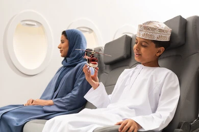 قضاء اوقات عائلية ممتعة في رحلة جويه ,مفهوم السياحة و السفر, رحلة سياحية من خلال خطوط الطيران العماني, صورة من الجانب لولد عربي خليجي عماني يرتدي الدشداشة و الكمه يجلس في مقعد الطائرة  بجانب والدته ويلعب بمجسم صغير لطائرة، ايماءات وجه تدل على السعادة