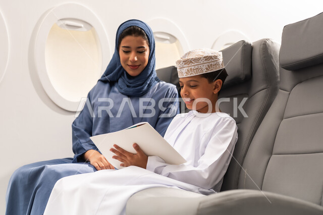 التعلم والابداع وتنمية المهارات, مفهوم السياحة و السفر, التنقل باستخدام وسائل النقل الجوية, ولد عربية خليجي عماني يرتدي الدشداشة و الكمه يجلس على مقعده داخل الطائرة برفقه والدته يمسك كراسة التلوين و القلم , ايماءات السعادة و السرور