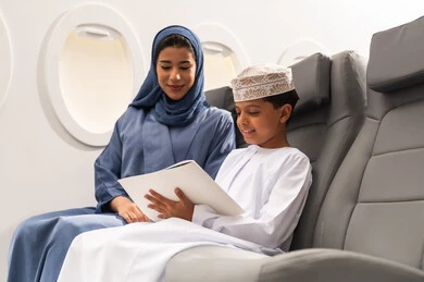 Integration and focus in drawing and coloring, the concept of tourism and travel, mobility using air transportation, an Omani Gulf Arab boy wearing a dishdasha and a kumma sitting in his seat inside the airplane with his mother, holding a coloring book and a pencil, gestures of happiness and joy.