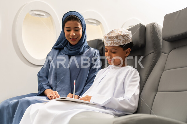 الاستمتاع بالرسم والتلوين, مفهوم السياحة و السفر, التنقل باستخدام وسائل النقل الجوية, ولد عربية خليجي عماني يرتدي الدشداشة و الكمه يجلس على مقعده داخل الطائرة برفقه والدته يمسك كراسة التلوين و القلم , ايماءات السعادة و السرور