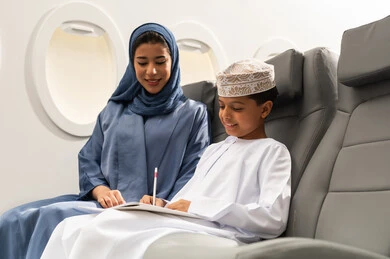 الاستمتاع بالرسم والتلوين, مفهوم السياحة و السفر, التنقل باستخدام وسائل النقل الجوية, ولد عربية خليجي عماني يرتدي الدشداشة و الكمه يجلس على مقعده داخل الطائرة برفقه والدته يمسك كراسة التلوين و القلم , ايماءات السعادة و السرور