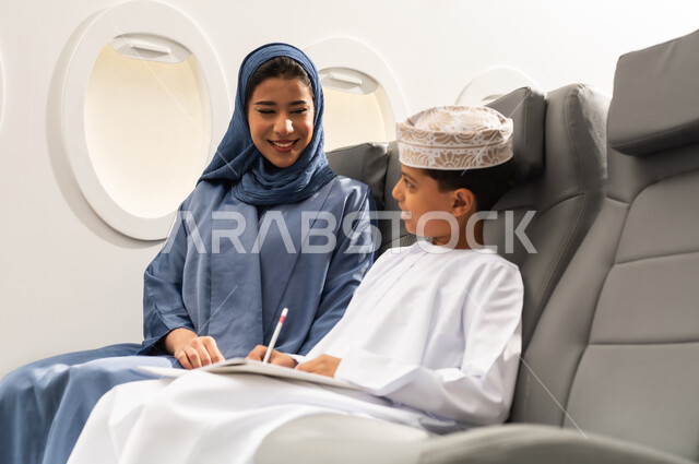 التعبير بالرسم عن الحب والمشاعر الإيجابية, مفهوم السياحة و السفر, التنقل باستخدام وسائل النقل الجوية, ولد عربية خليجي عماني يرتدي الدشداشة و الكمه يجلس على مقعده داخل الطائرة برفقه والدته يمسك كراسة التلوين و القلم , ايماءات السعادة و السرور