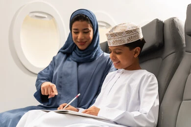 الاندماج والتركيز في الرسم والتلوين, مفهوم السياحة و السفر, التنقل باستخدام وسائل النقل الجوية, ولد عربية خليجي عماني يرتدي الدشداشة و الكمه يجلس على مقعده داخل الطائرة برفقه والدته يمسك كراسة التلوين و القلم , ايماءات السعادة و السرور