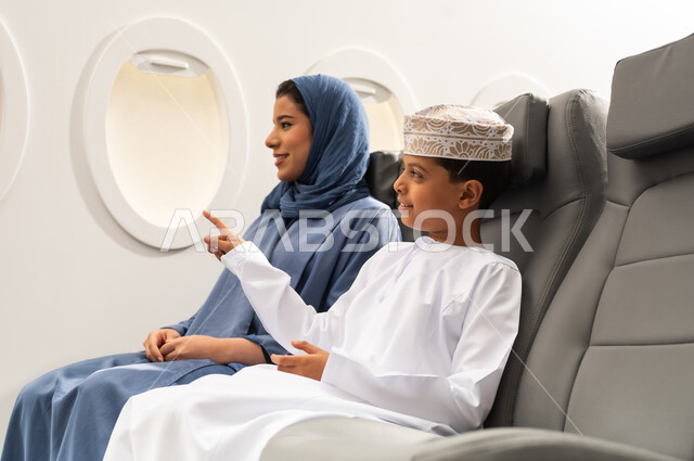 وسائل النقل الجوي والرحلات حول العالم, الاشارة الى شي ما, مفهوم السياحة و السفر, ولد عربي خليجي عماني يرتدي الدشداشة و الكمة يجلس على مقعده داخل الطائرة برفقه والدته , ايماءات السعادة و السرور