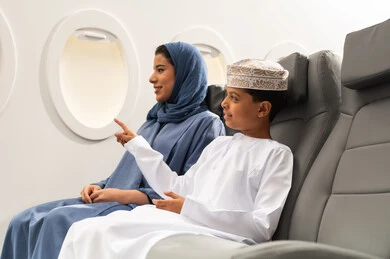 وسائل النقل الجوي والرحلات حول العالم, الاشارة الى شي ما, مفهوم السياحة و السفر, ولد عربي خليجي عماني يرتدي الدشداشة و الكمة يجلس على مقعده داخل الطائرة برفقه والدته , ايماءات السعادة و السرور