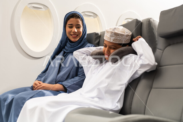 The feeling of laziness and lethargy, a family tourist trip, an Omani Arab woman wearing a hijab and abaya with her son inside the airplane, the feeling of tiredness and exhaustion and the need for sleep and rest, spending the summer vacation with family, Oman Air.