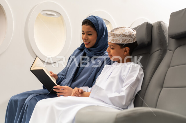 ايماءات التركيز والاستمتاع, قراءة الروايات والقصص، مفهوم السياحة و السفر, التنقل باستخدام وسائل النقل الجوية, امراة عربيه خليجيه عمانية ترتدي الحجاب و العباءة تجلس بجانب ابنها داخل الطائرة و تمسك كتاب