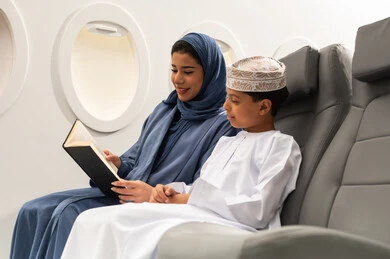 ايماءات التركيز والاستمتاع, قراءة الروايات والقصص، مفهوم السياحة و السفر, التنقل باستخدام وسائل النقل الجوية, امراة عربيه خليجيه عمانية ترتدي الحجاب و العباءة تجلس بجانب ابنها داخل الطائرة و تمسك كتاب