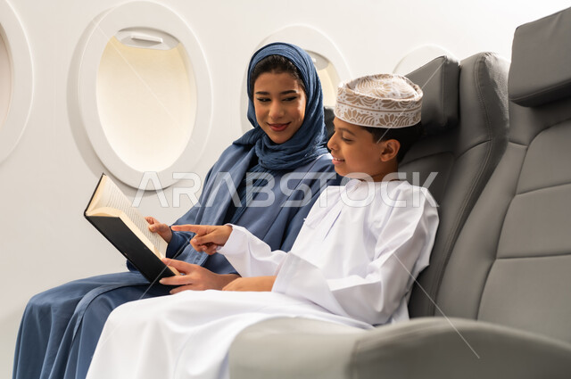قراءة الروايات والقصص، ايماءات التركيز والاستمتاع،مفهوم السياحة و السفر, التنقل باستخدام وسائل النقل الجوية, امراة عربيه خليجيه عمانية ترتدي الحجاب و العباءة تجلس بجانب ابنها داخل الطائرة و تمسك كتاب