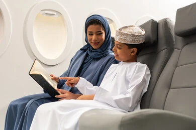 Reading novels and stories, gestures of focus and enjoyment, the concept of tourism and travel, moving using air transportation, an Arab Gulf Omani woman wearing a hijab and abaya sitting next to her son inside the airplane holding a book.