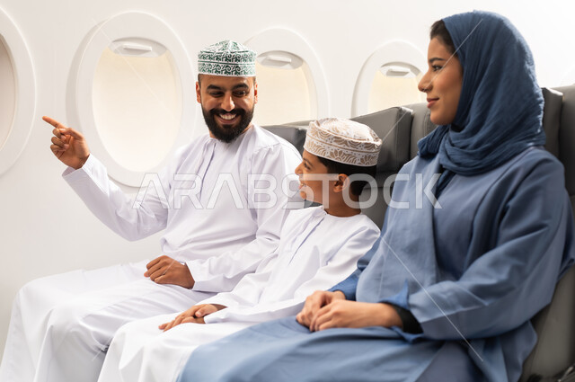 قضاء وقت ممتع داخل الطائرة , عائله عربيه خليجيه عمانية ترتدي الزي التقليدي تجلس على المقعد داخل الطائرة فى رحله جويه , ايماءات السعاده و السرور , رجل خليجي يقوم بالاشارة الى شيء ما
