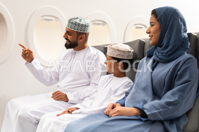 الانتقال عبر الطائرات الحديثة , الاستعداد لقضاء عطله صيفية ممتعه ,عائلة عربيه خليجيه عمانية ترتدي الزي التقليدي تجلس داخل الطائرة بايماءات السعاده و السرور , رجل عربي خليجي يرتدي الدشداشة و الكمه يقوم بالاشارة الي شيء ما