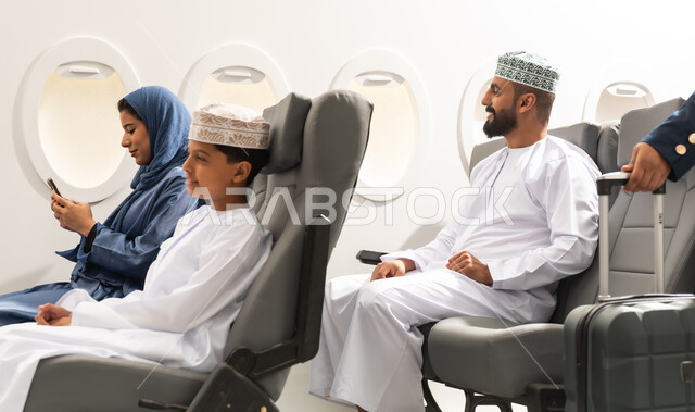 السفر بين المدن والدول, عائلة عربيه خليجيه عمانية ترتدي الزي التقليدي تجلس داخل الطائرة بايماءات السعاده و السرور , امراة عربيه خليجيه تمسك الهاتف المحمول , تصفح مواقع التواصل الالكتروني