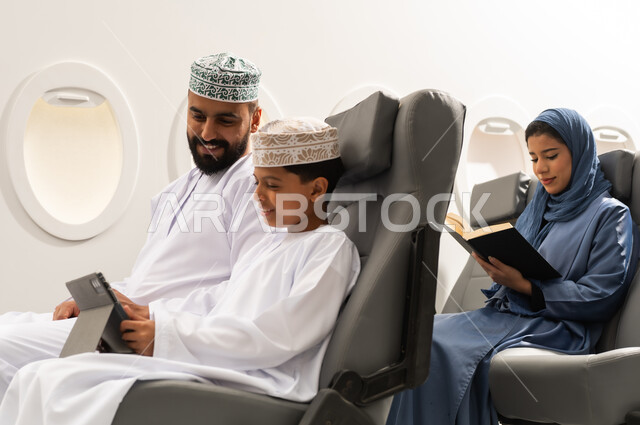 مشاهدة الأنشطة الترفيهية والتعليمية، دمج التعلم بالتقنية والتكنولوجيا, امراة خليجيه تقوم بقراءة كتاب, مفهوم السياحه و السفر , ولد عربية خليجي عماني يرتدي الدشادشة و الكمة يجلس داخل الطائرة فى رحله جوية برفقة والده يمسك بيده جهاز التابلت, ايماءات السعادة و السرور