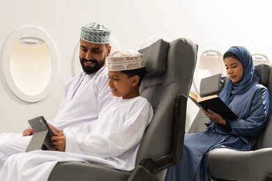 Watching recreational and educational activities, integrating learning with technology and tech, a Gulf woman reading a book, the concept of tourism and travel, a Gulf Omani boy wearing a dishdasha and kumma sitting inside an airplane on a flight with his father holding a tablet in his hand, gestures of happiness and joy.