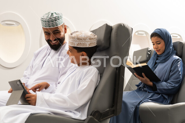 دمج التعلم بالتقنية والتكنولوجيا, امراة خليجيه تقوم بقراءة كتاب، اهتمام الآباء بالأبناء، مشاهدة الأنشطة الترفيهية والتعليمية, مفهوم السياحه و السفر , ولد عربية خليجي عماني يرتدي الدشادشة و الكمة يجلس داخل الطائرة فى رحله جوية برفقة والده يمسك بيده جهاز التابلت, ايماءات السعادة و السرور