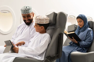 دمج التعلم بالتقنية والتكنولوجيا, امراة خليجيه تقوم بقراءة كتاب، اهتمام الآباء بالأبناء، مشاهدة الأنشطة الترفيهية والتعليمية, مفهوم السياحه و السفر , ولد عربية خليجي عماني يرتدي الدشادشة و الكمة يجلس داخل الطائرة فى رحله جوية برفقة والده يمسك بيده جهاز التابلت, ايماءات السعادة و السرور