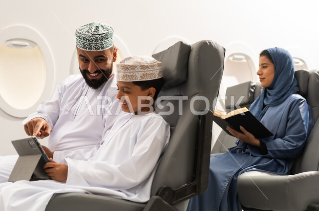 A Gulf woman is reading a book, watching recreational and educational activities, the concept of tourism and travel, an Omani Gulf Arab boy wearing a dishdasha and a kumma sitting inside the airplane on a flight with his father holding a tablet in his hand, gestures of happiness and joy.