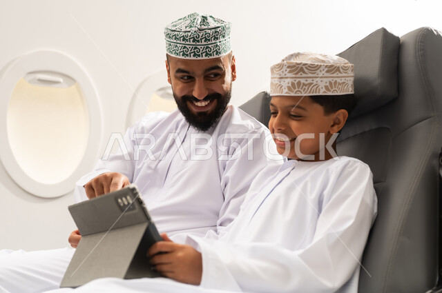 اهتمام الآباء بالأبناء، متابعة منصات التعليم الالكتروني عبر الانترنت، دمج التعلم بالتقنية والتكنولوجيا، مشاهدة الأنشطة الترفيهية والتعليمية, مفهوم السياحه و السفر , ولد عربية خليجي عماني يرتدي الدشادشة و الكمة يجلس داخل الطائرة فى رحله جوية برفقة والده يمسك بيده جهاز التابلت, ايماءات السعادة و السرور