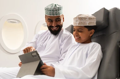 Parents' interest in their children, following online educational platforms, integrating learning with technology, watching recreational and educational activities, the concept of tourism and travel, an Omani Gulf Arab boy wearing a dishdasha and kumma sitting inside an airplane on a flight with his father holding a tablet in his hand, gestures of happiness and joy.