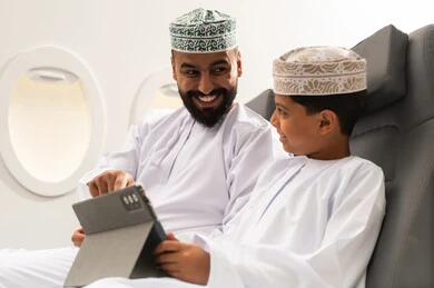 متابعة منصات التعليم الالكتروني عبر الانترنت، اهتمام الآباء بالأبناء، دمج التعلم بالتقنية والتكنولوجيا، مشاهدة الأنشطة الترفيهية والتعليمية, مفهوم السياحه و السفر , ولد عربية خليجي عماني يرتدي الدشادشة و الكمة يجلس داخل الطائرة فى رحله جوية برفقة والده يمسك بيده جهاز التابلت, ايماءات السعادة و السرور